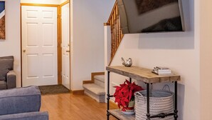 A modern living room with a flat-screen TV mounted on the wall, a wooden console table with decorative items, a staircase, and a door leading to another room. - Spacious Mountain Home with Private Hot Tub (Granby)