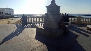Une place agrémentée d’un monument central, de bancs et d’une balustrade donnant sur une étendue d’eau.
