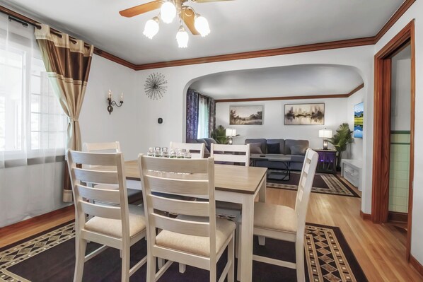 A dining area with a wooden table and chairs, a ceiling fan, and a view of the living room.