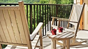 A balcony with two wooden chairs and a table, a black railing, and a view of a forested hill.