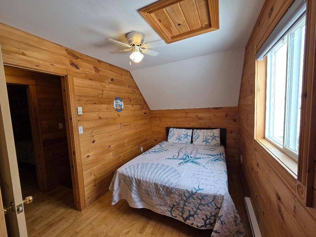 Ein kleiner, gemütlicher Raum mit Holzwänden, einem Einzelbett mit gemustertem Bettbezug, einem Deckenventilator und einem Fenster, das natürliches Licht hereinlässt.