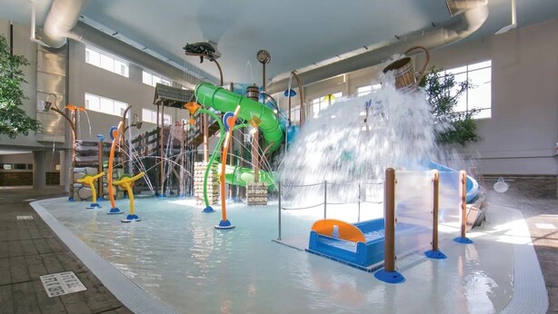 An indoor water park with a green slide, multiple water jets, and a blue water play area.