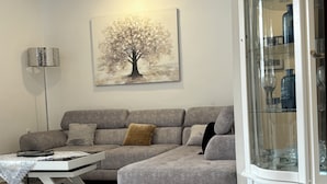 A living room with a grey sofa, a coffee table, a painting of a tree, and a cabinet with glass doors. - A rare pearl (Oran)
