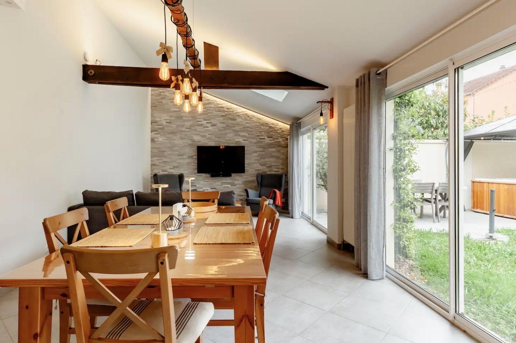 Un espace repas moderne avec une longue table et des chaises en bois, un mur en pierre agrémenté d’un téléviseur à écran plat, et de grandes portes-fenêtres coulissantes donnant sur un jardin.