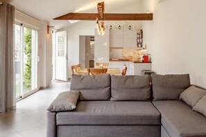 A modern living room with a grey sectional sofa, a wooden ceiling beam, and a kitchen in the background.