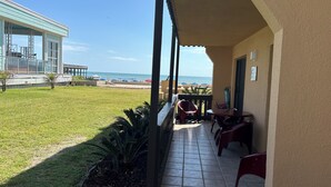 En overdækket terrasse med udsigt til stranden, et lille bord og stole, samt en bygning med glasfacade.