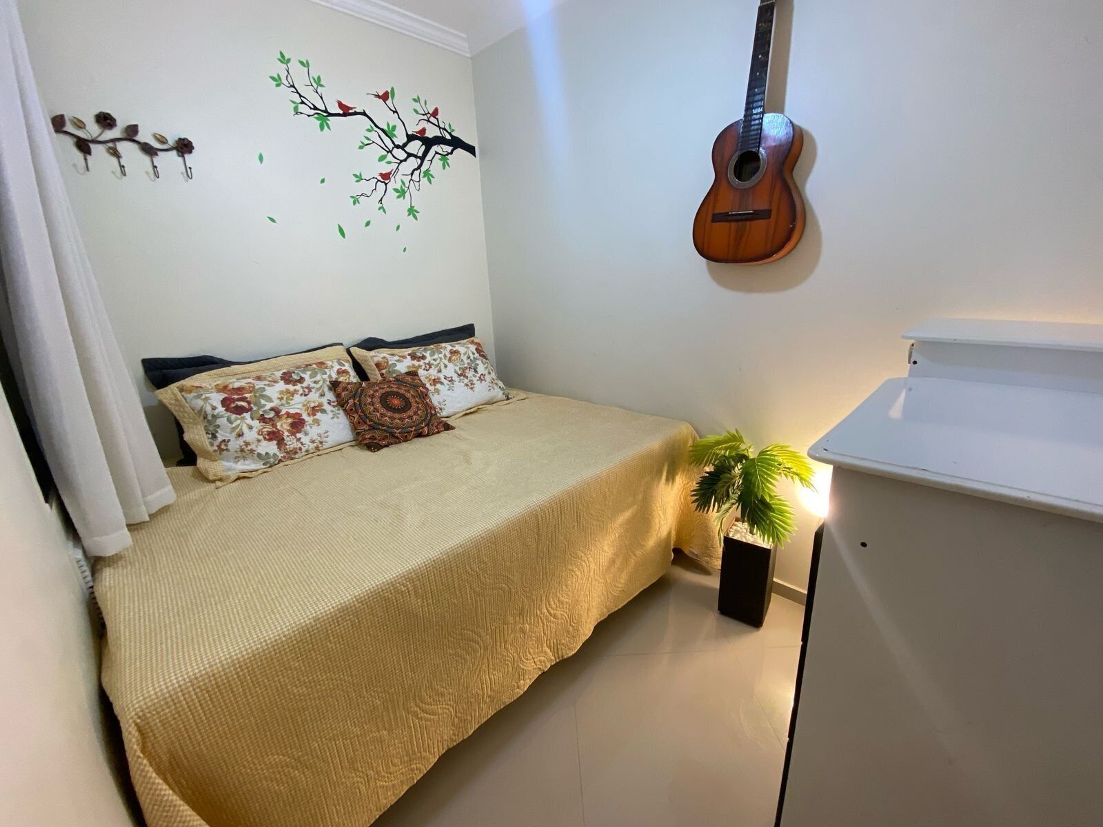 A bedroom with a bed, a wall-mounted guitar, a small potted plant, and a white cabinet.