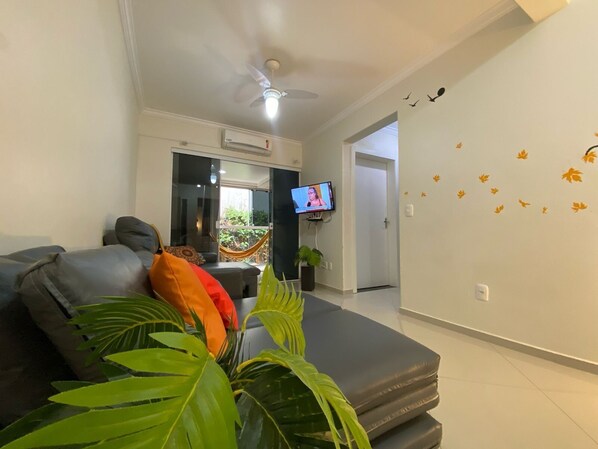 A living room with a sofa, a television, and a ceiling fan. - Apto 203 Ed Dambros no centro (Piratuba)