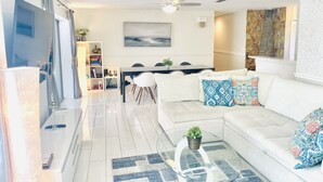 A modern living room with a white sofa, a glass coffee table, and a dining area with a table and chairs.