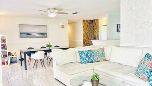 A modern living room with a white sofa, colorful cushions, a glass coffee table, and a dining area with a black table and chairs.