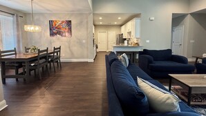 A spacious living room with a dark blue sofa, a dining area with a wooden table and chairs, and a kitchen in the background. - Spacious 3BR Auburn Getaway – Family-Ready and Near All Auburn Has to Offer (Auburn)