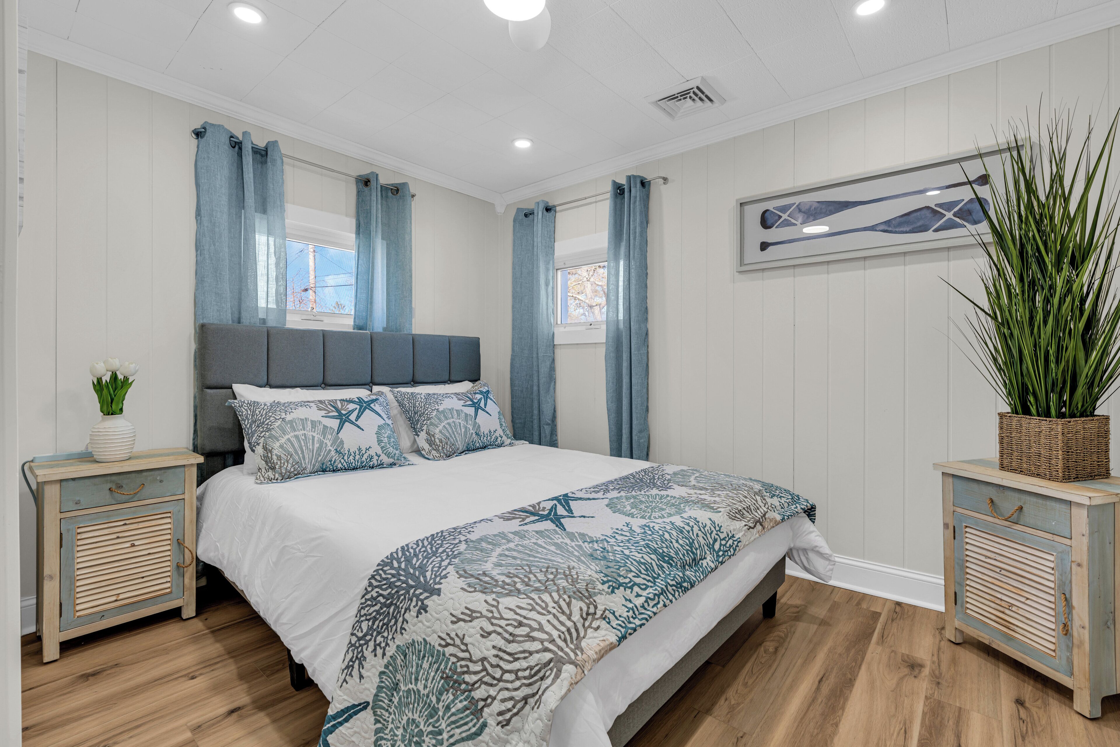 Un dormitorio con cama, cortinas, mesita de noche, planta en maceta y un cuadro colgado en la pared.