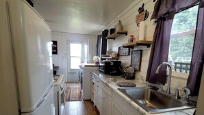 A kitchen with white cabinets, a stainless steel sink, and a refrigerator. There are wooden floors and a window with curtains. - Comfortable and Cozy 1-bedroom house in delightful Rapid City with AC & Wifi (Rapid City)