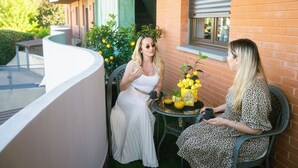 Two women sitting at a table with a 'welcome' sign and lemons, on a balcony with a brick wall and greenery in the background.