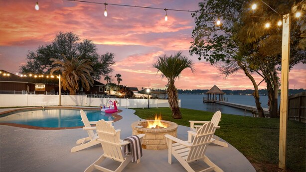 Un espace extérieur avec une piscine, un foyer et des fauteuils Adirondack, entouré d’arbres et offrant une vue sur un plan d’eau au coucher du soleil.