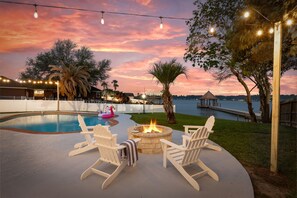 An outdoor pool area with a fire pit and Adirondack chairs, surrounded by trees and a view of a body of water at sunset. - Pool Fun on Lake Conroe - Gameroom - Paddleboards (Willis)