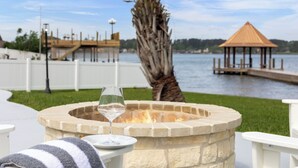 Un foyer en pierre, un verre de vin blanc, un palmier et un gazebo en bois au bord d’un lac.