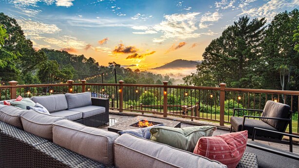 Une terrasse dotée de mobilier d’extérieur, d’un foyer et offrant une vue sur le coucher de soleil au-dessus d’une zone boisée.