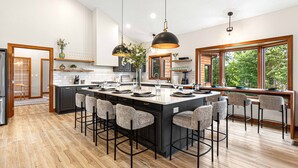 Une cuisine moderne dotée d’un îlot central, de tabourets de bar, de luminaires suspendus et de grandes fenêtres offrant une vue sur la verdure.