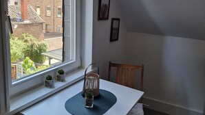 Ein Zimmer mit einem weißen Schreibtisch, einem Stuhl, einem Fenster mit Blick auf Gebäude und einer kleinen Pflanze auf der Fensterbank.