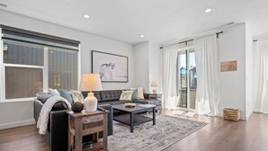 A modern living room with a black sofa, a coffee table, and a rug. There is a large window with curtains, a framed picture on the wall, and a small side table with a lamp. - Bear Cub’s Den R5983 | Snowbasin Ski Escape | EV (Mountain Green)