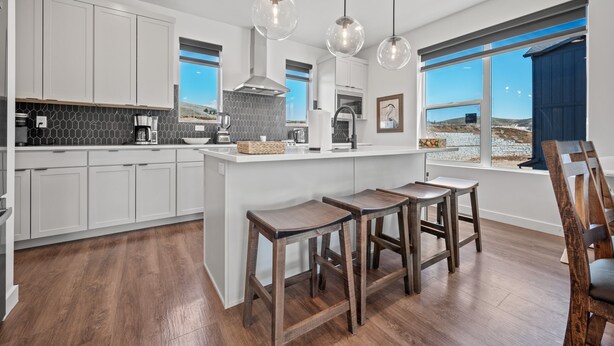 A modern kitchen with white cabinets, a central island with stools, a coffee maker, and a view of the outdoors through large windows. - Bear Cub’s Den R5983 | Snowbasin Ski Escape | EV (Mountain Green)