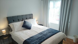 Un lit bien fait, avec une tête de lit sombre, une literie blanche et un plaid bleu foncé. On y trouve une table de chevet avec une lampe, ainsi qu’une fenêtre avec des rideaux.