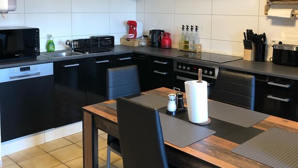 A modern kitchen with white cabinets, a black countertop, and a wooden dining table with chairs. - 2 room apartment with terrace (Roth-sur-Our)