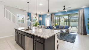 A modern kitchen with granite countertops, a dishwasher, and a central island. There is a staircase with white railings leading to an upper level. A living area with a sofa and a coffee table is visible, with a large window offering a view of the outdoors. - 5BR Poolside Retreat (Kissimmee)