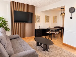A living room with a sofa, a coffee table, a flat-screen TV mounted on a wooden panel, a dining area with a table and chairs, and a kitchen area with a stove.