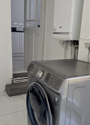 Eine Waschmaschine mit Frontlader in einem Waschraum, inklusive digitaler Bedienkonsole, einer Tür zu einem Flur und einem weißen, wandmontierten Warmwasserbereiter.