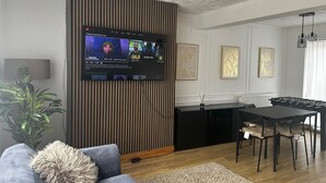 Een moderne woonkamer met een flatscreen televisie bevestigd aan een houten paneelwand, een grijze bank, een klein vloerkleed en een eethoek met een zwarte tafel en stoelen.