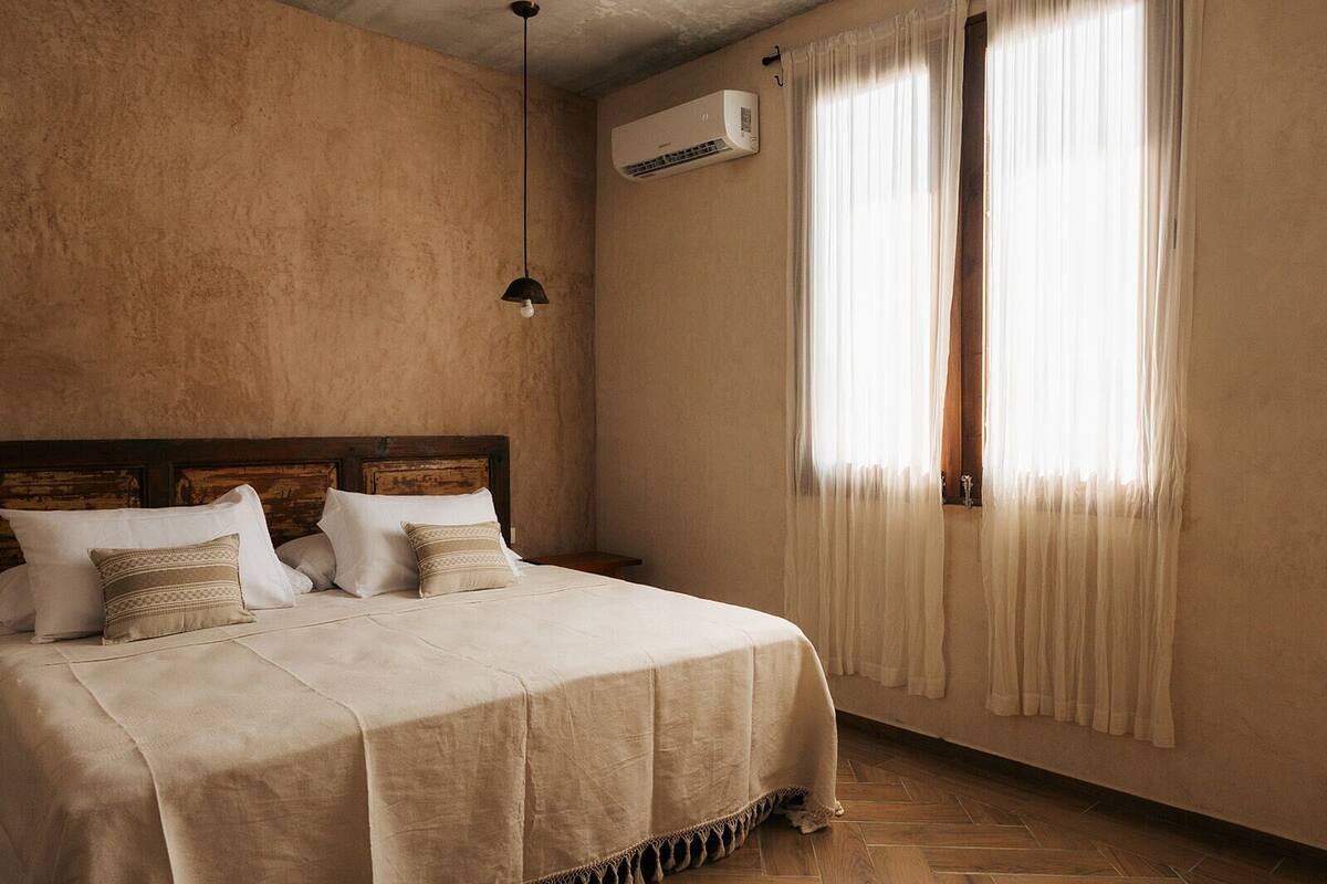 A bedroom with a wooden headboard, a bed with a beige bedspread and pillows, a hanging pendant light, an air conditioning unit, and a window with sheer curtains.