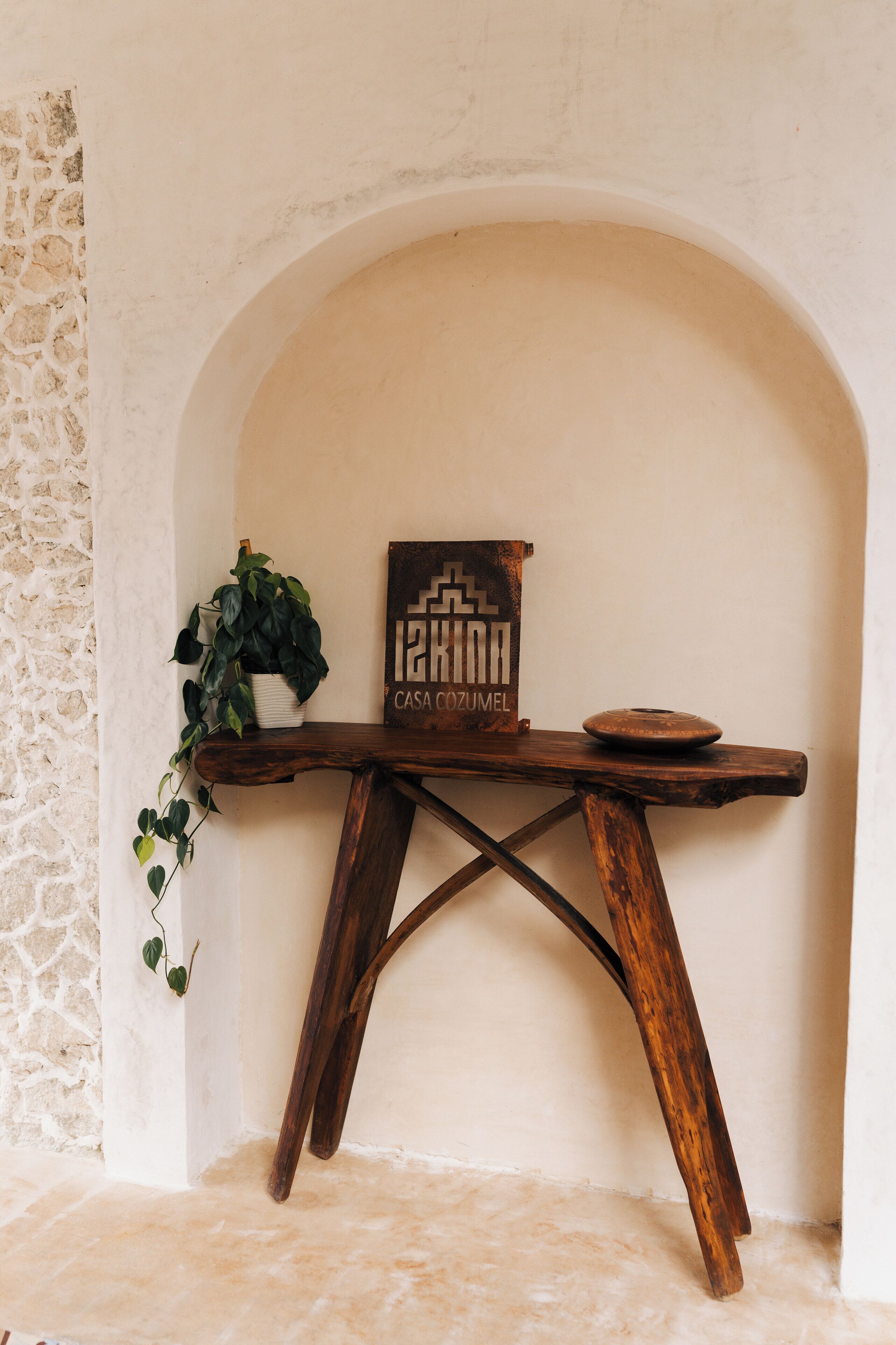 A rustic wooden table with a potted plant and a decorative piece against a white wall with an arched doorway.
