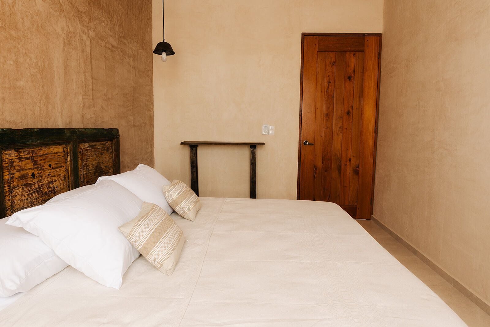 Un dormitorio con cama, almohadas, cabecera de madera, una mesita, una puerta y una lámpara colgante.