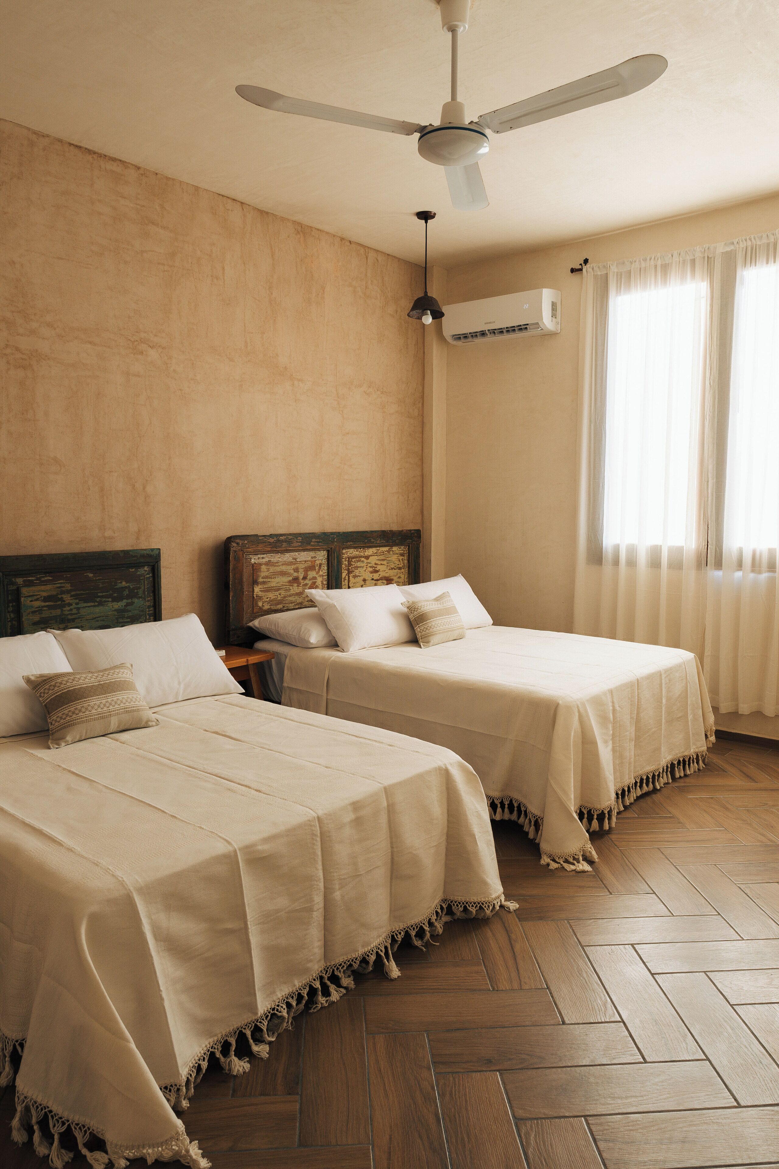 Habitación de hotel con dos camas, ventilador de techo, unidad de aire acondicionado y una ventana con cortinas.