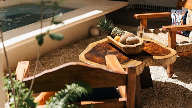 A cozy outdoor seating area with wooden furniture, a small table with fruits and a glass, and a view of a bedroom through an open door. - Casa Hotel Izkina (Cozumel)