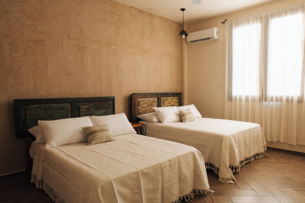 A room with two beds, a wooden floor, an air conditioning unit, and a window with curtains.