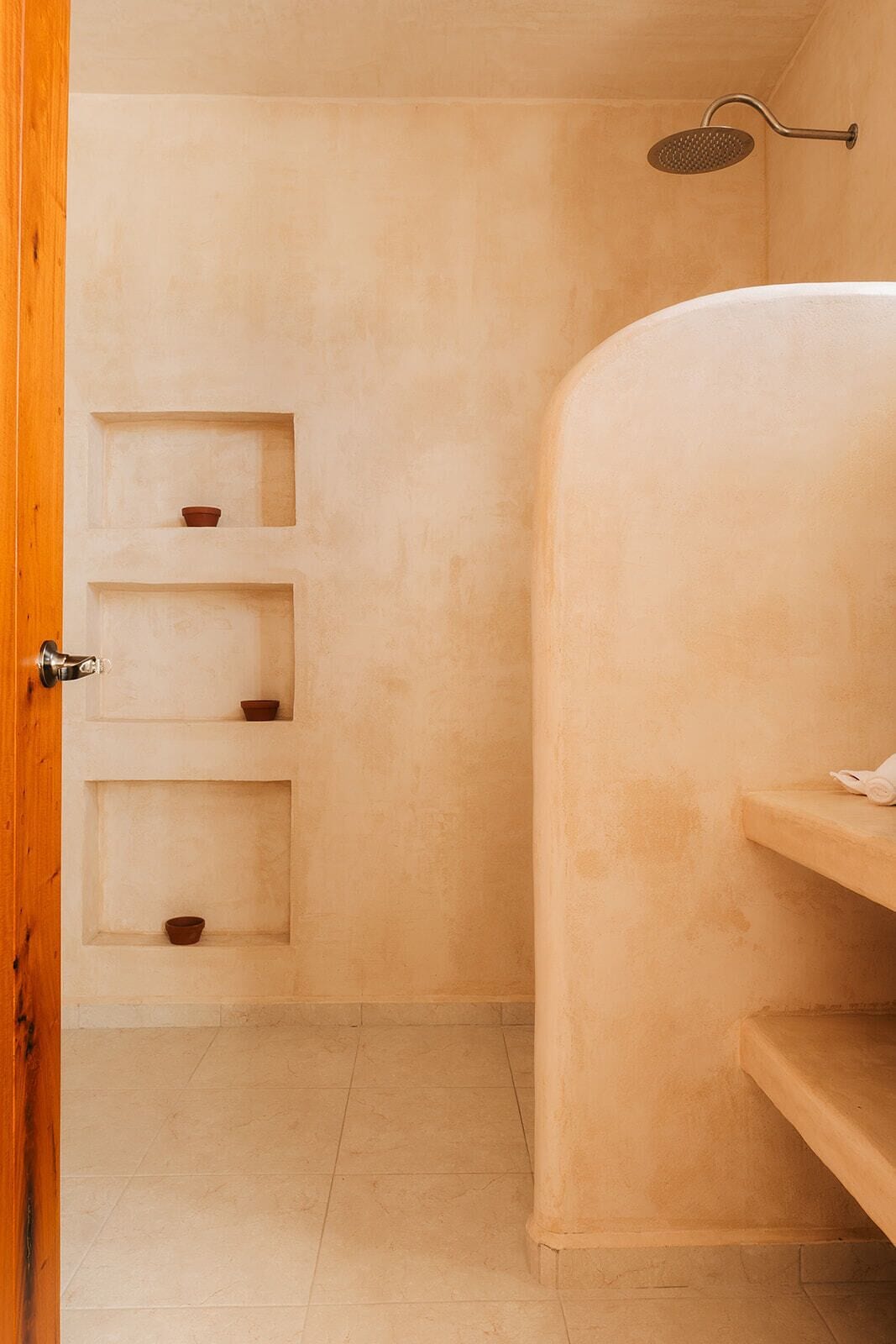 A bathroom with a wooden door, a shower with a handheld showerhead, and three shelves with small bowls.