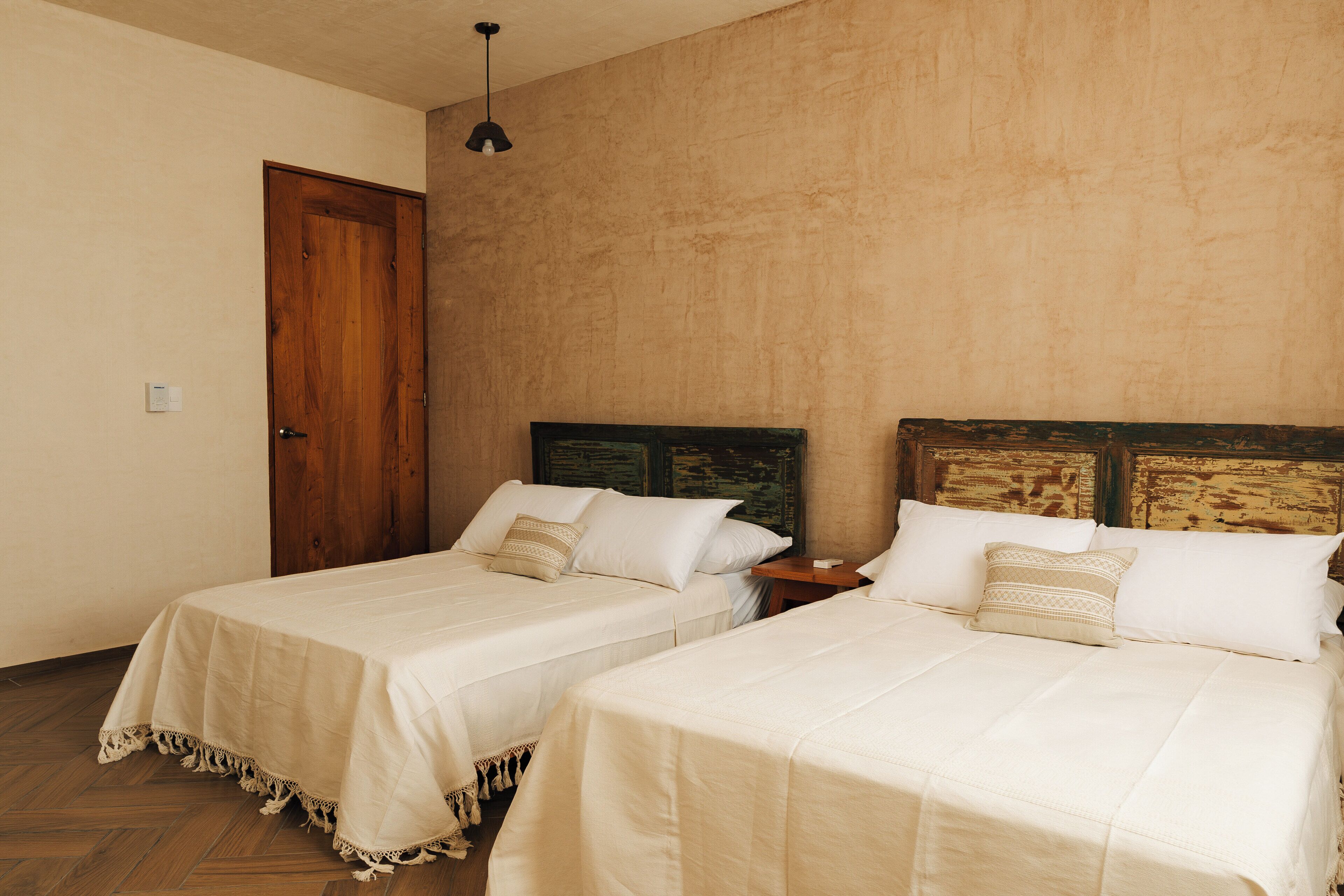 A room with two beds, wooden headboards, a wooden door, and a small table with a lamp.