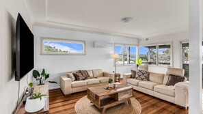 A living room with a flat-screen TV, two sofas, a coffee table, and large windows offering a view of the outdoors. - Blue Pacific Holiday Home - Murrah St 73 (Bermagui)