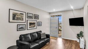 A living room with a black leather sofa, a flat-screen TV mounted on the wall, and a door leading to an outdoor area.