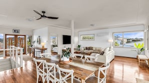 A spacious living area with a wooden dining table, white chairs, a sofa, and a television.