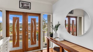 A room with a wooden door, a mirror, a white wall, and a view of the outside.