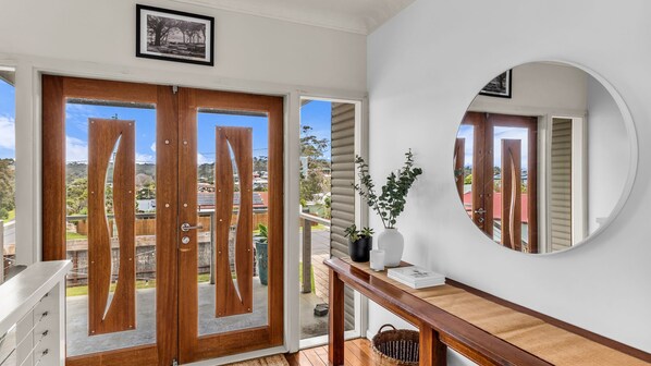A room with a wooden door, a mirror, a white wall, and a view of the outside. - Blue Pacific Holiday Home - Murrah St 73 (Bermagui)