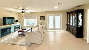 A spacious living room with a sofa, coffee table, and TV. There is a large window and a ceiling fan.
