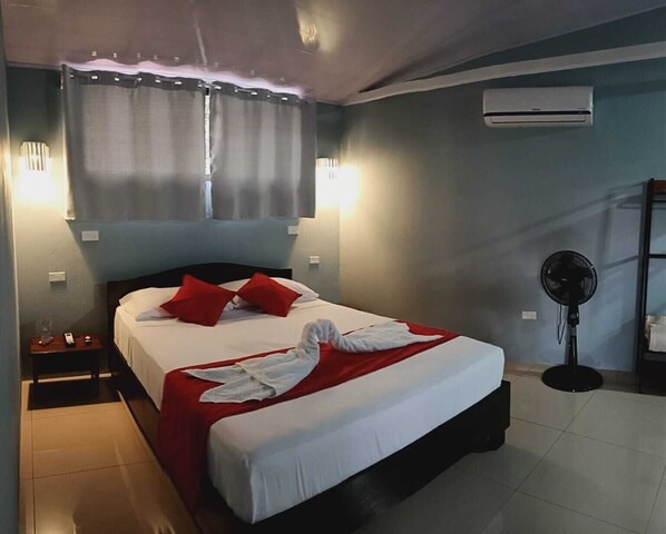 Une chambre d’hôtel avec un lit, des oreillers rouges, une couverture blanche, une serviette, un climatiseur, un ventilateur sur pied et une table de chevet avec une lampe et un téléphone.