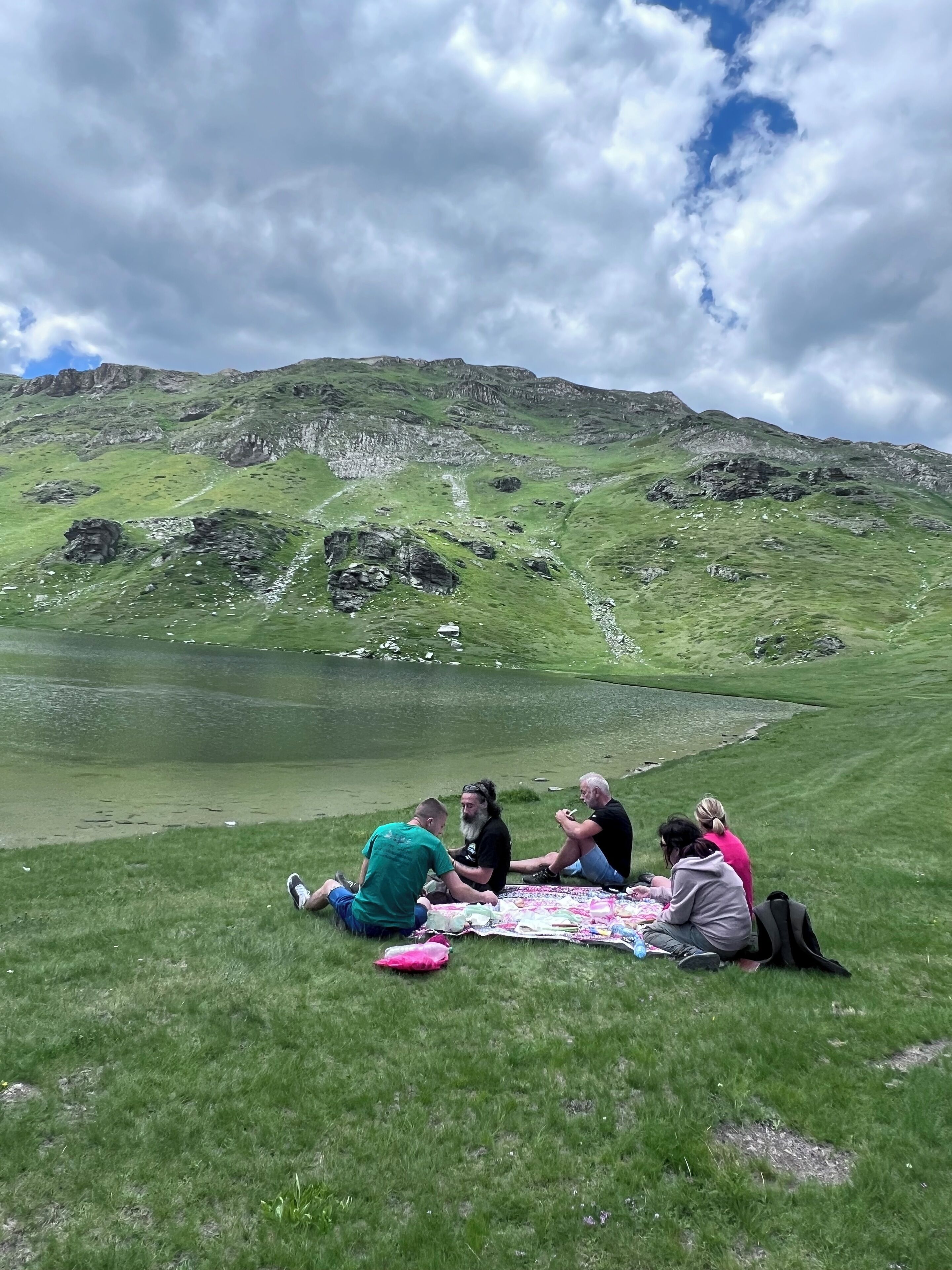 Un grupo de personas sentadas sobre una manta junto a un lago, con montañas al fondo.