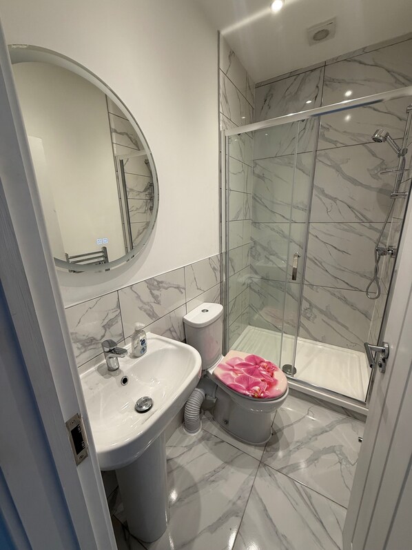 Un baño moderno con cabina de ducha en mosaico de mármol, un espejo redondo y un inodoro con asiento de color rosa adornado con una orquídea.