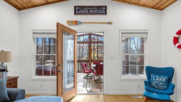 Une pièce avec un plafond en bois, une pancarte indiquant « bienvenue à la MAISON DU LAC », une pagaie, une chaise bleue avec un coussin et une vue sur une terrasse extérieure avec des chaises rouges.